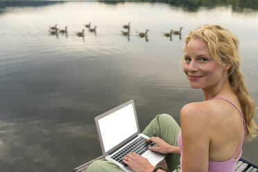 Young woman using laptop on a jetty at a lake - JOSF03610