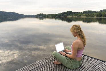 Young woman using laptop on a jetty at a lake - JOSF03611