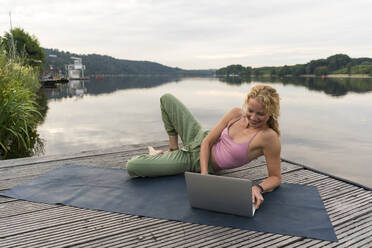 Smiling young woman using laptop on a jetty at a lake - JOSF03612