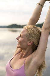 Happy young woman raising her arms at a lake - JOSF03618