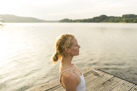 Junge Frau mit geschlossenen Augen auf einem Steg an einem See, lizenzfreies Stockfoto