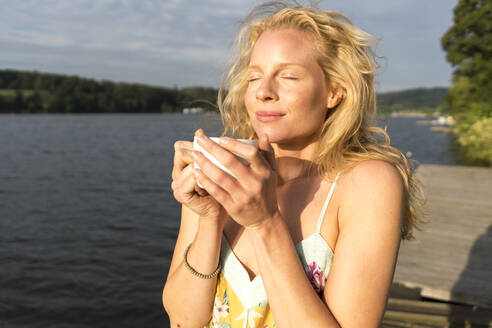 Young woman dwith closed eyes holding a cup at a lake - JOSF03639