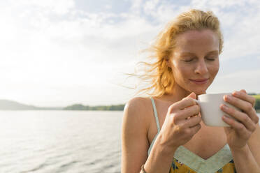 Junge Frau mit geschlossenen Augen hält eine Tasse an einem See - JOSF03640