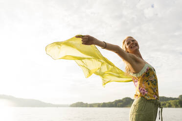 Happy young woman holding a cloth at a lake - JOSF03643