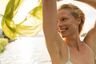 Smiling young woman holding a cloth at a lake - JOSF03644