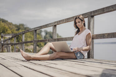 Woman sitting on wood bridge, using laptop - JOSF03703