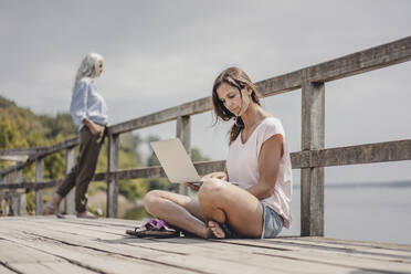 Woman sitting on wood bridge, using laptop, mother standing in background - JOSF03704