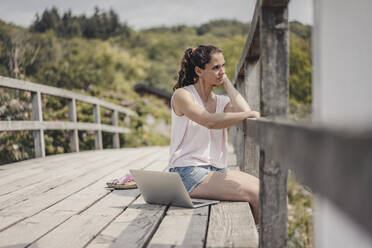 Frau mit Laptop auf einer Holzbrücke sitzend, mit Blick auf die Aussicht - JOSF03706