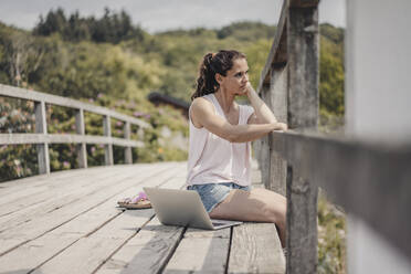 Woman with laptop sitting on wood bridge, looking at view - JOSF03706