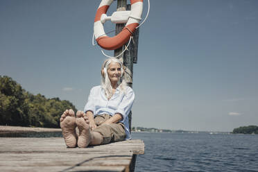 Senior woman sitting on jetty, listening music by the sea - JOSF03714