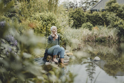Senior woman sitting at garden pond, listening music - JOSF03720