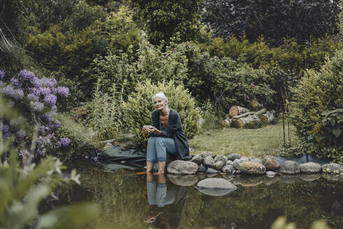 Woman relaxing at garden pond, drinking tea - JOSF03722