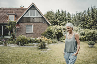 Ältere Frau, die in ihrem Garten spazieren geht, mit den Händen in den Taschen - JOSF03779