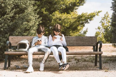 Zwei Kinder in Astronauten- und Superheldenkostümen benutzen ein Mobiltelefon auf einer Parkbank, lizenzfreies Stockfoto
