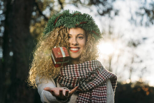 Porträt einer lächelnden jungen Frau mit Weihnachtskranz auf dem Kopf, die mit einem Weihnachtsgeschenk jongliert - DAMF00058