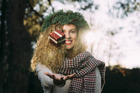 Porträt einer lächelnden jungen Frau mit Weihnachtskranz auf dem Kopf, die mit einem Weihnachtsgeschenk jongliert, lizenzfreies Stockfoto