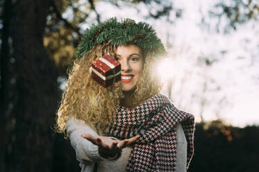 Porträt einer lächelnden jungen Frau mit Weihnachtskranz auf dem Kopf, die mit einem Weihnachtsgeschenk jongliert - DAMF00059