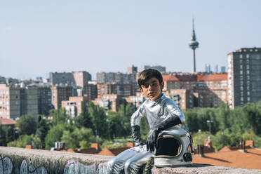 Portrait of boy dressed as an astronaut sitting on a wall in the city - JCMF00214