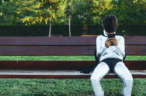 Portrait of boy disguised as superhero using mobile phone on park bench stock photo