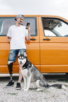 Junger Mann mit Hund, der eine Beinprothese trägt und ein Mobiltelefon im Wohnmobil benutzt - CJMF00017