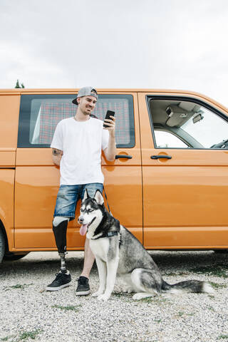 Junger Mann mit Hund, der eine Beinprothese trägt und ein Mobiltelefon im Wohnmobil benutzt, lizenzfreies Stockfoto