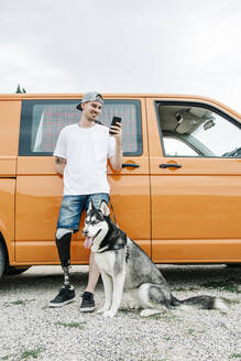 Junger Mann mit Hund, der eine Beinprothese trägt und ein Mobiltelefon im Wohnmobil benutzt - CJMF00018