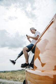 Junger Mann mit Beinprothese sitzt auf dem Dach eines Wohnmobils und benutzt ein Mobiltelefon - CJMF00019