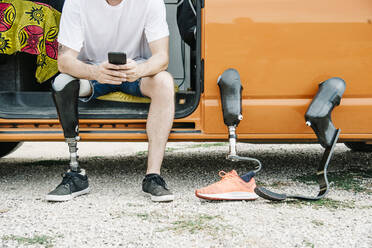 Young man with leg prosthesis sitting in camper van using cell phone - CJMF00021