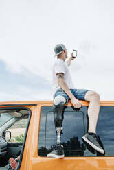 Young man with leg prosthesis sitting on roof of camper van using cell phone - CJMF00023