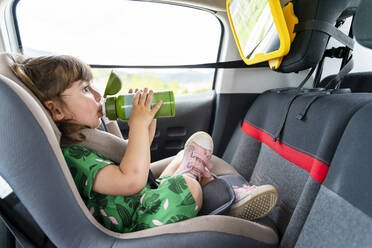 Toddler girl sitting on a car seat with a mirror drinking water - GEMF03149