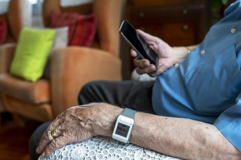 Älterer Mann, der zu Hause ein Mobiltelefon benutzt und ein intelligentes Notfallalarmarmband um das Handgelenk trägt - GEMF03151