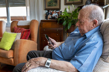 Älterer Mann, der zu Hause ein Mobiltelefon benutzt und ein intelligentes Notfallalarmarmband um das Handgelenk trägt - GEMF03152
