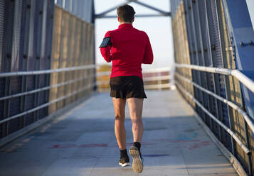 Rear view of a jogger with smartphone in arm pocket running on a bridge - JSRF00582