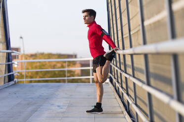 Jogger with smartphone in arm pocket, stretching his leg on a bridge railing - JSRF00584