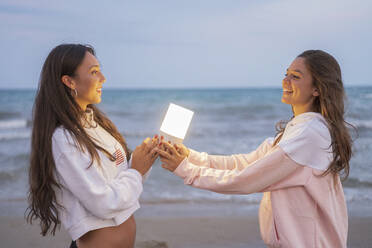 Two happy female friends holding led light on the beach at dusk - DLTSF00124