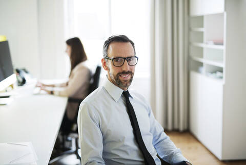 Porträt eines selbstbewussten Geschäftsmannes im Büro mit einem Angestellten im Hintergrund, lizenzfreies Stockfoto