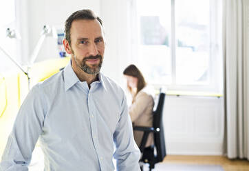 Portrait of confident businessman in office with employee in background - MIKF00015