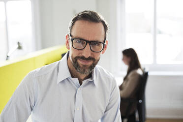 Porträt eines selbstbewussten Geschäftsmannes im Büro mit einem Angestellten im Hintergrund - MIKF00018