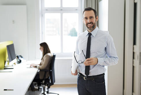 Porträt eines lächelnden Geschäftsmannes im Büro mit Angestellten im Hintergrund - MIKF00021
