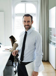 Portrait of smiling businessman in office with employee in background - MIKF00022