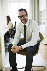 Portrait of smiling businessman in office with employee in background - MIKF00027