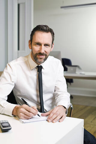 Porträt eines selbstbewussten Geschäftsmannes, der im Büro am Schreibtisch sitzt und Notizen macht, lizenzfreies Stockfoto