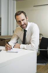 Porträt eines selbstbewussten Geschäftsmannes, der im Büro am Schreibtisch sitzt und Notizen macht - MIK00059