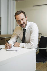 Portrait of confident businessman sitting at desk in office taking notes - MIK00059