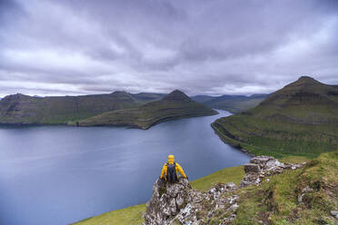 Wanderer auf Felsen sitzend mit Blick auf die Fjorde, Funningur, Insel Eysturoy, Färöer Inseln, Dänemark, Europa - RHPLF12101