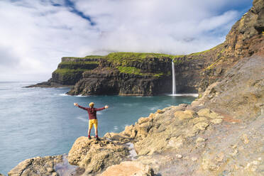 Man on cliffs with open arms admiring Gasadalur waterfall, Vagar island, Faroe Islands, Denmark, Europe - RHPLF12128