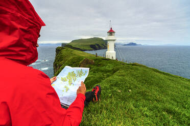 Hiker on cliffs looks at the map next to lighthouse, Mykines island, Faroe Islands, Denmark, Europe - RHPLF12134