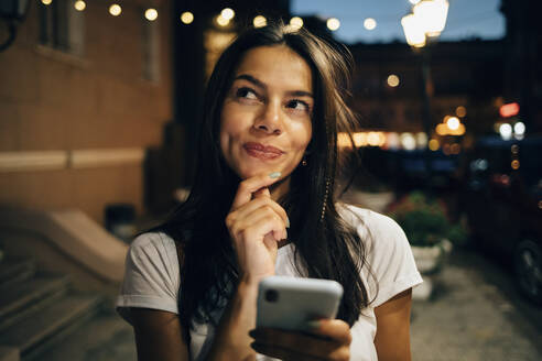 Young woman using smartphone in the city at night, looking up - OYF00078