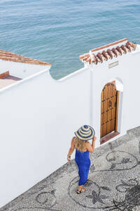 Rückenansicht einer Frau in blauem Kleid und Strohhut, die eine Gasse entlanggeht, Nerja, Spanien - LJF00990