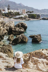 Rückenansicht einer Frau in weißem Kleid und Strohhut, die auf einem Felsen sitzt und auf das Meer blickt, Nerja, Spanien - LJF00994